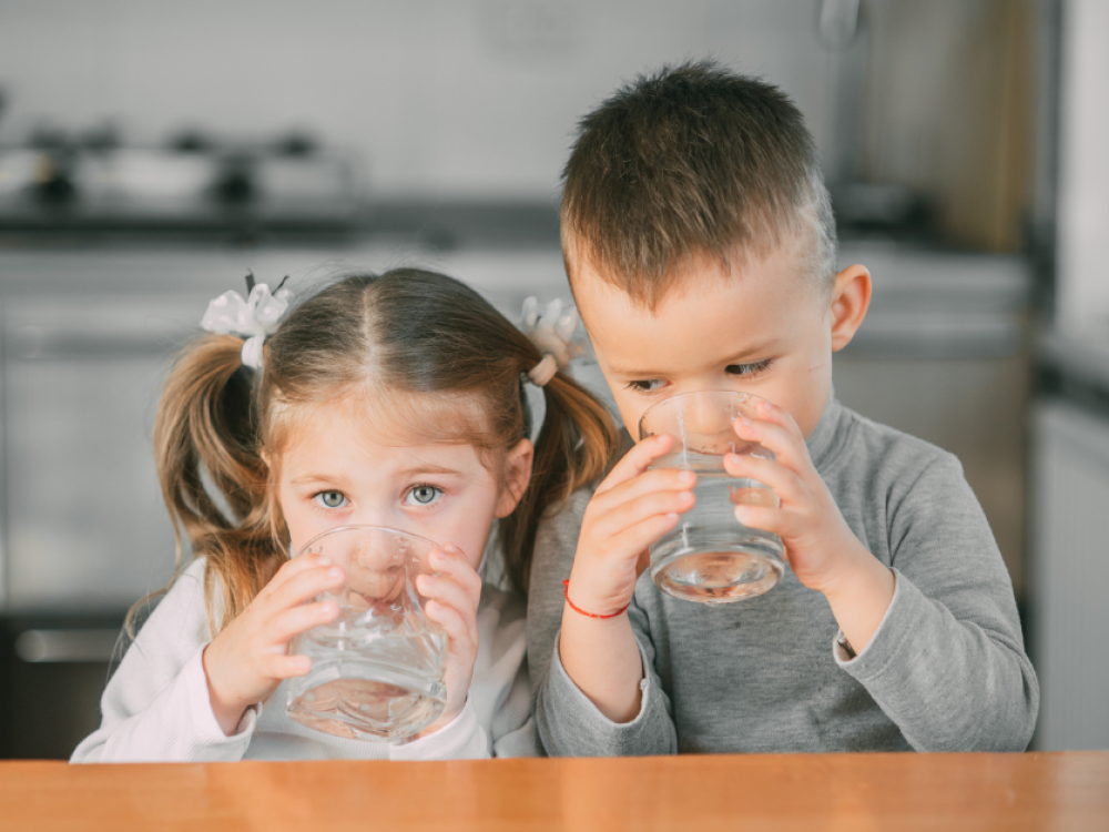 Two young children sitting at a table, drinking water from clear glasses, with a blurred kitchen background.